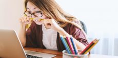 Woman biting yellow pencil in her mouth in front of a computer