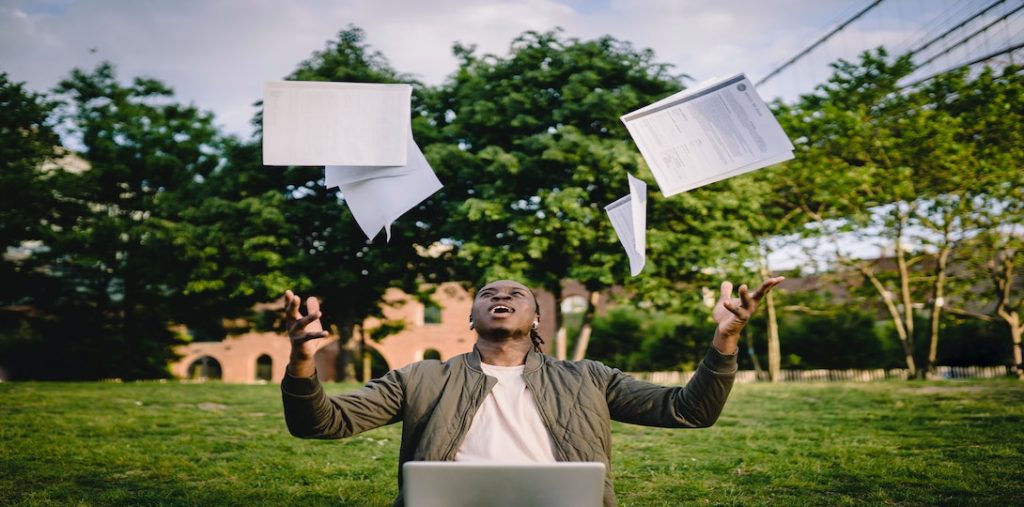 Young man at computer tosses papers in relief; one of the online teaching habits is to take a down day or day off