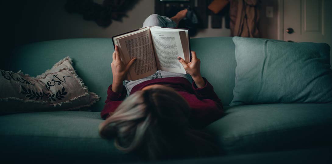 Woman on soft couch laying upside down, reading a book