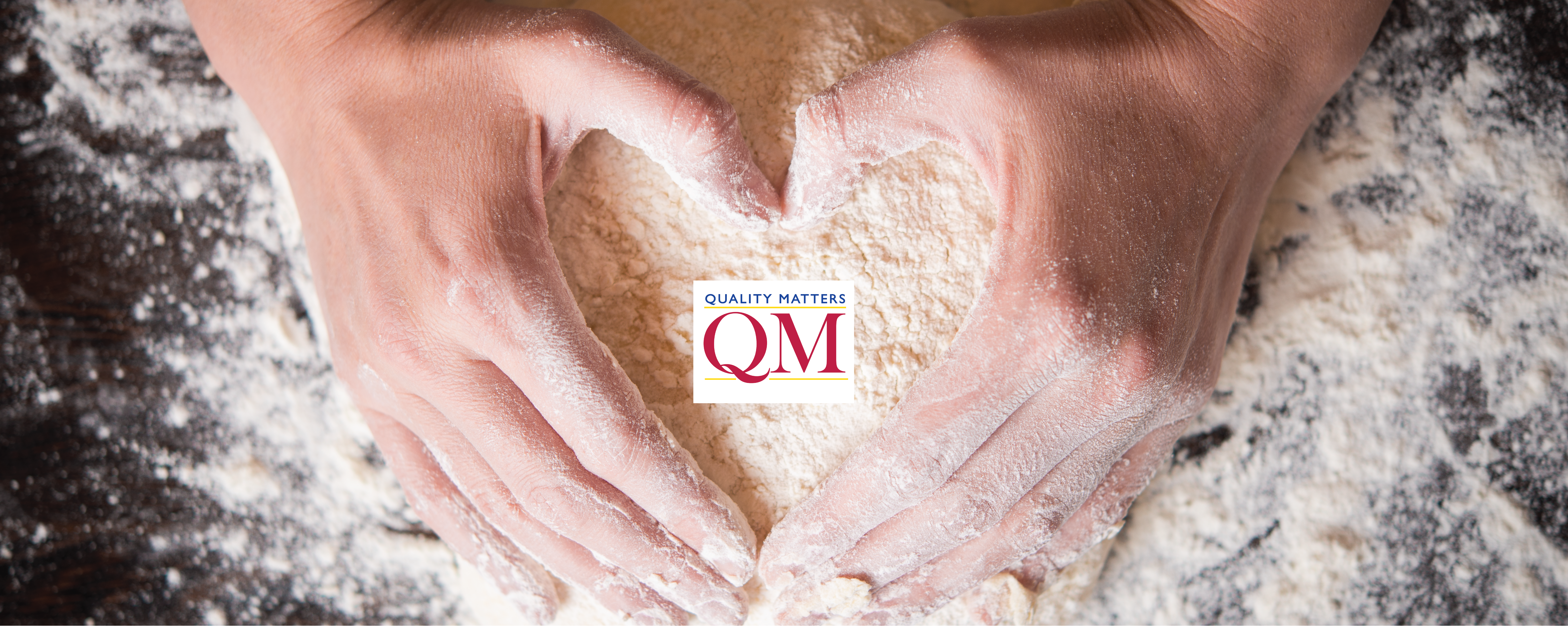 Two hands kneeding dough for bread. The hands are making the shape of a heart and the middle of the dough features a QM logo.